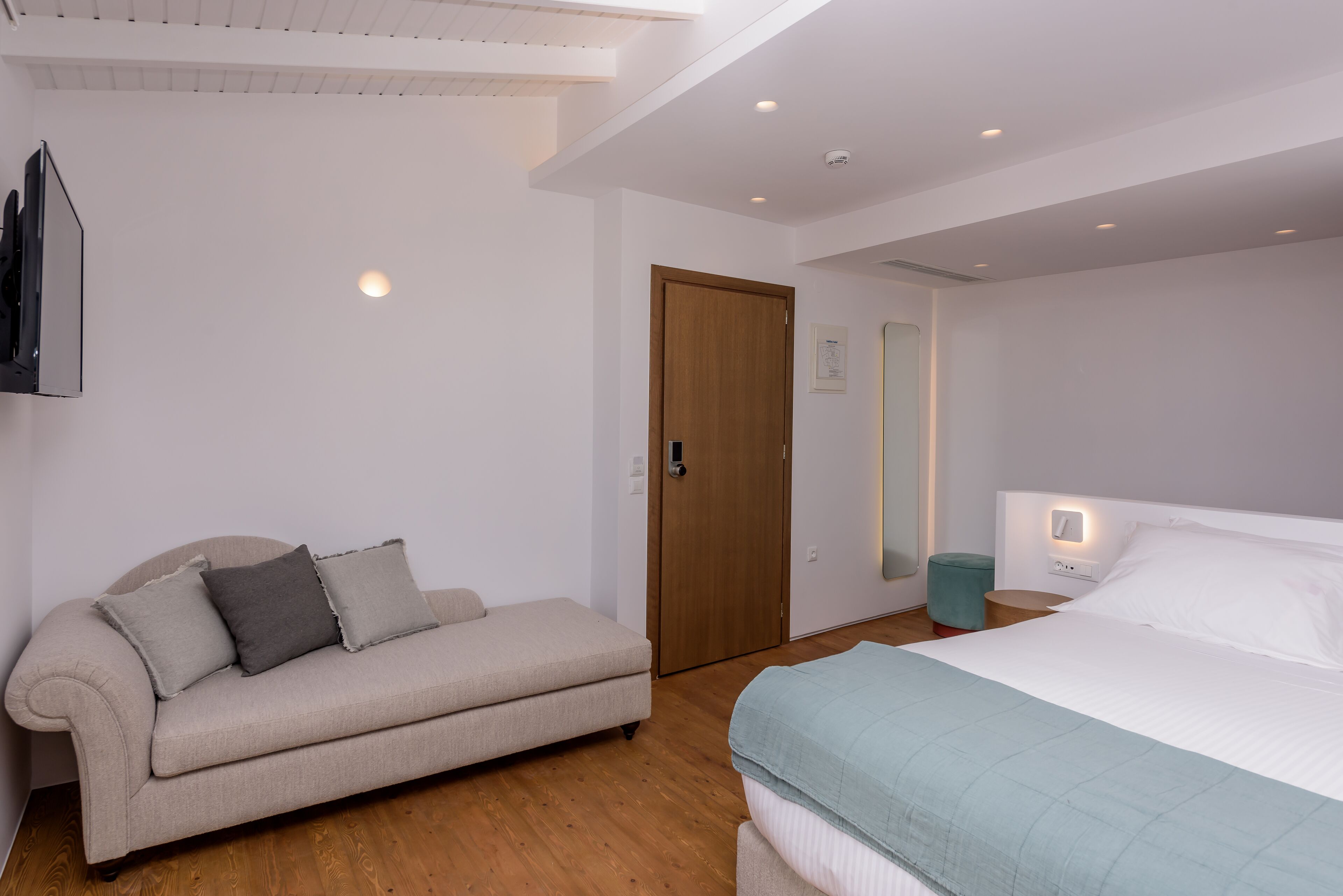A bedroom with a bed, a sofa, a wooden floor, a door, and a wall-mounted light fixture.