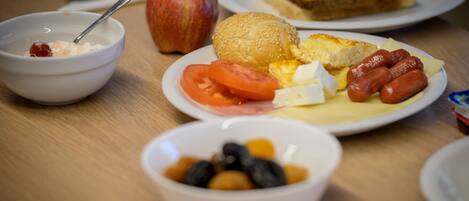 Una mesa de desayuno con un tazón de cereal, una manzana, un sándwich, un plato con huevos, tomates, queso y salchichas.