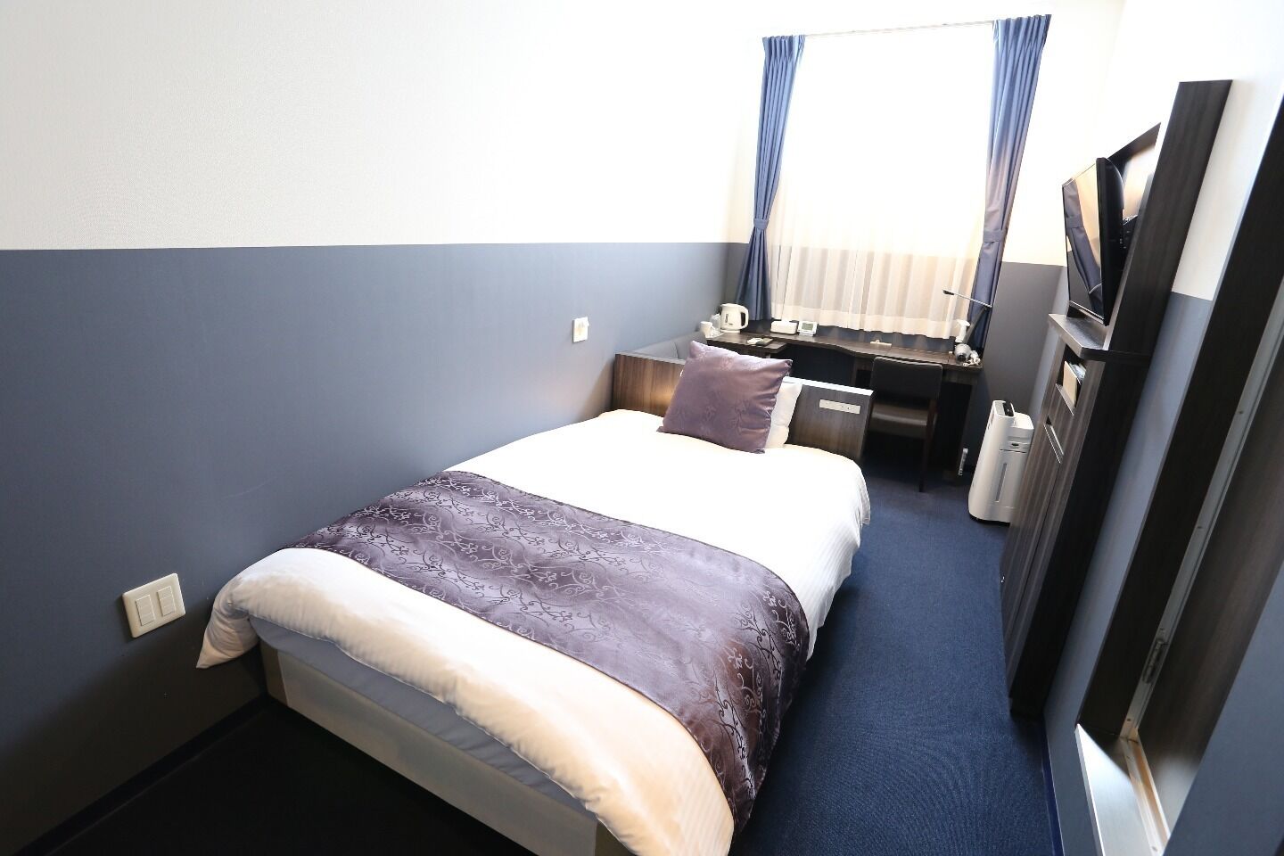 A hotel room with a bed, a desk, a chair, a TV, and a window with curtains.