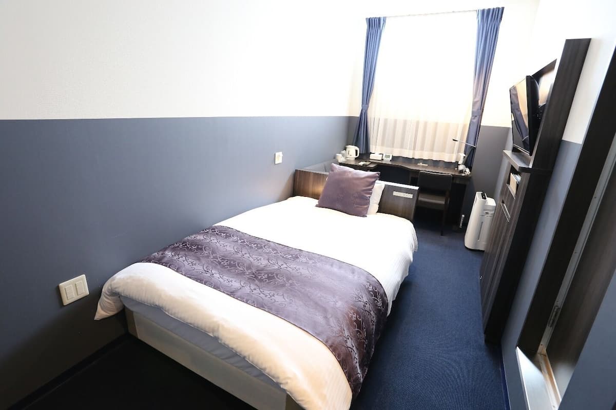 Una camera d'albergo con un letto, una scrivania, una sedia, una televisione e una finestra con tende.