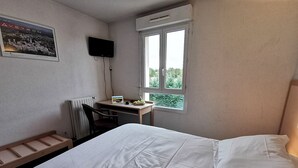 A hotel room with a bed, a desk with a chair, a radiator, a window with a view, and a TV mounted on the wall.
