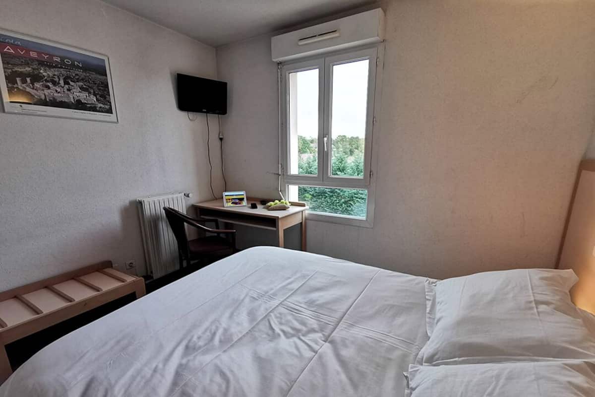 A hotel room with a bed, a desk with a chair, a radiator, a window with a view, and a TV mounted on the wall.