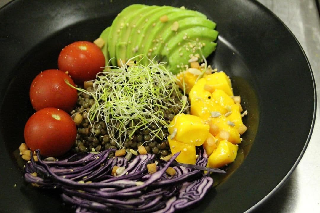 A black plate with a variety of food items including avocado, mango, red cabbage, and sprouts.