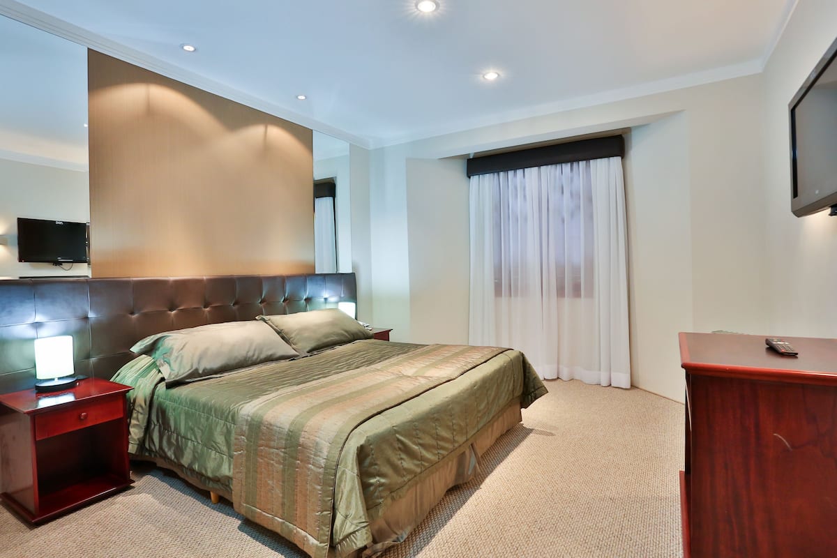 A hotel room with a large bed, a wooden nightstand, a desk, a television, and a window with curtains.