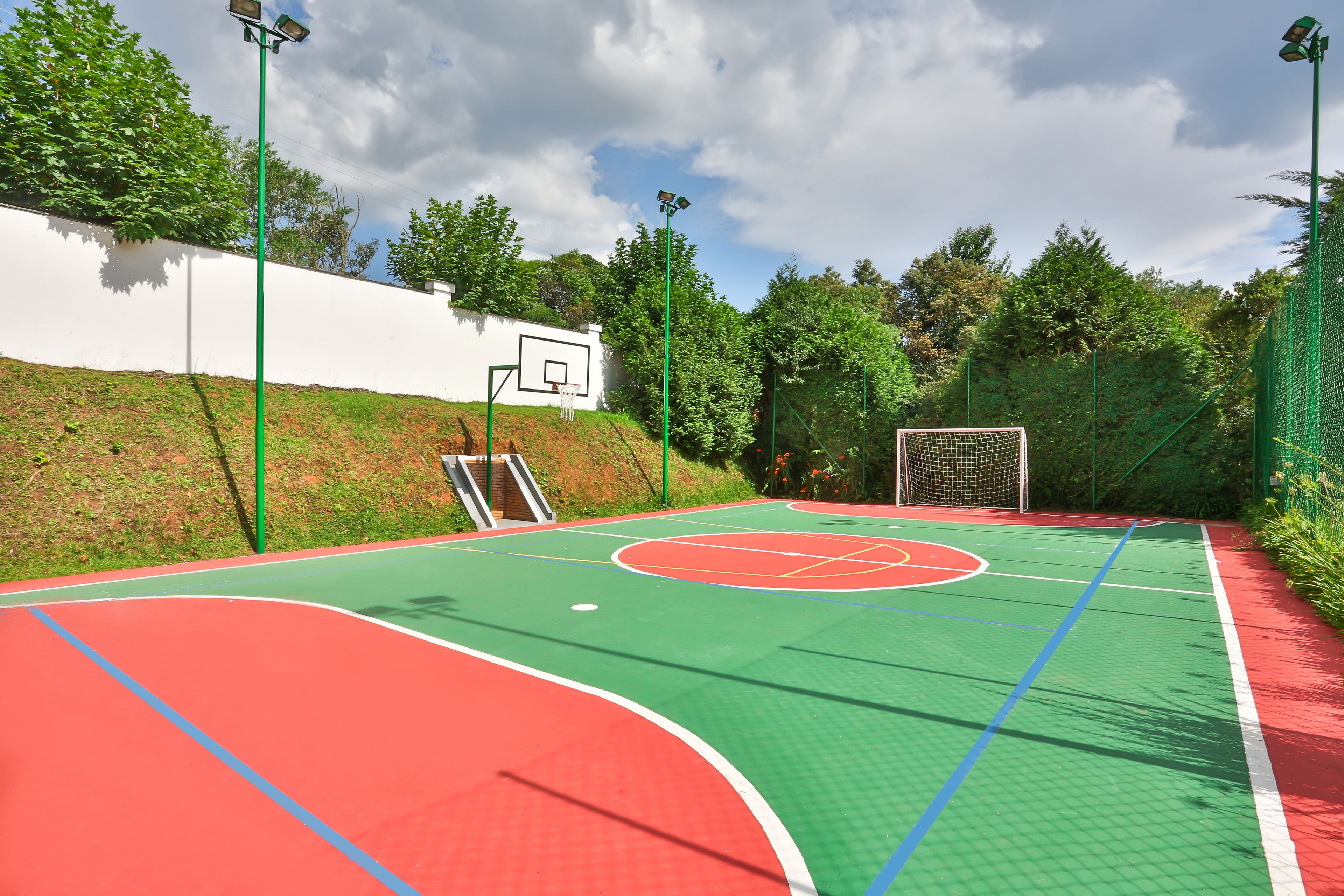 Un campo da basket all'aperto con superficie verde e rossa, un muro bianco e alberi sullo sfondo.