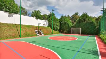 Un campo da basket all'aperto con superficie verde e rossa, un muro bianco e alberi sullo sfondo.