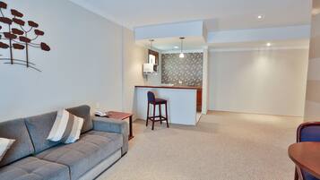 A modern living room with a grey sofa, a wooden table, and a kitchen area with a bar stool.