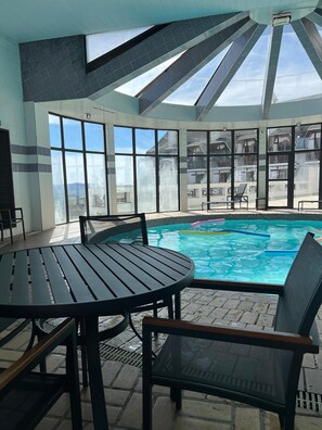 A sunlit indoor swimming pool area with a table and chairs, large windows, and a view of a building outside. - Home Green Home (Campos do Jordão)