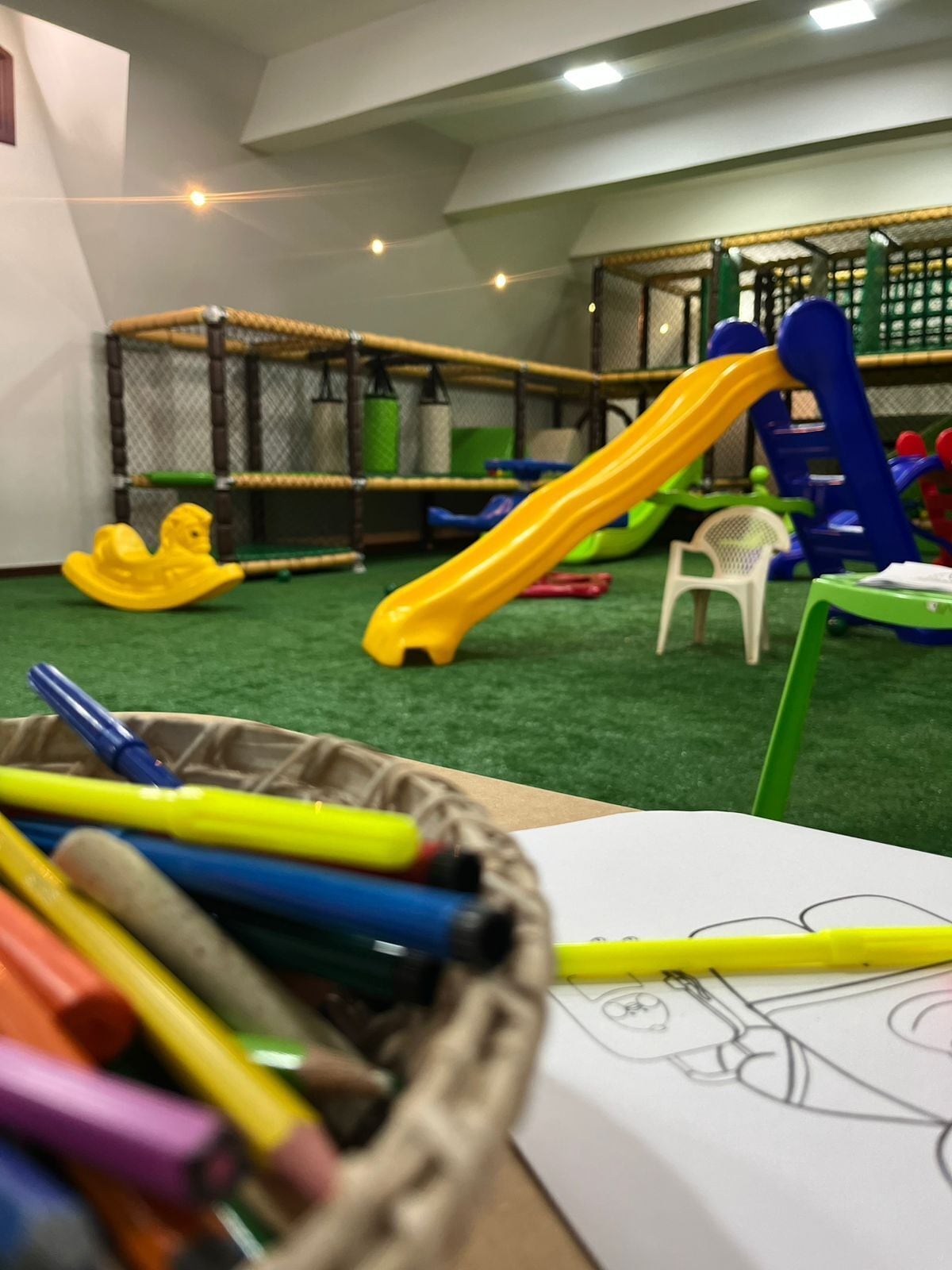 Zona infantil con toboganes y estructuras para escalar, un cesto de lápices de colores y un dibujo en papel.