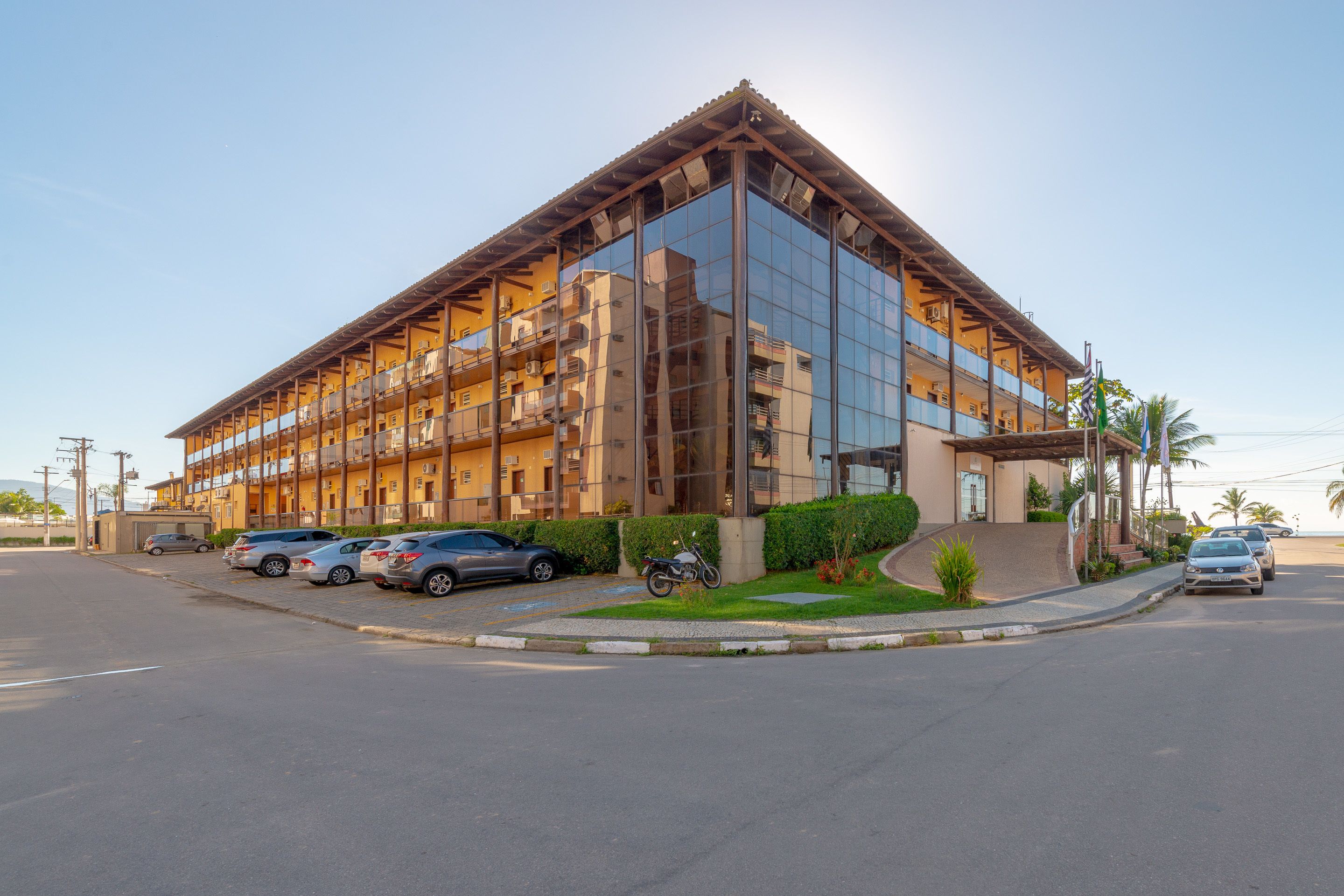A modern hotel with a glass facade, multiple balconies, and a parking area with several cars.