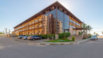 A modern hotel with a glass facade, multiple balconies, and a parking area with several cars.