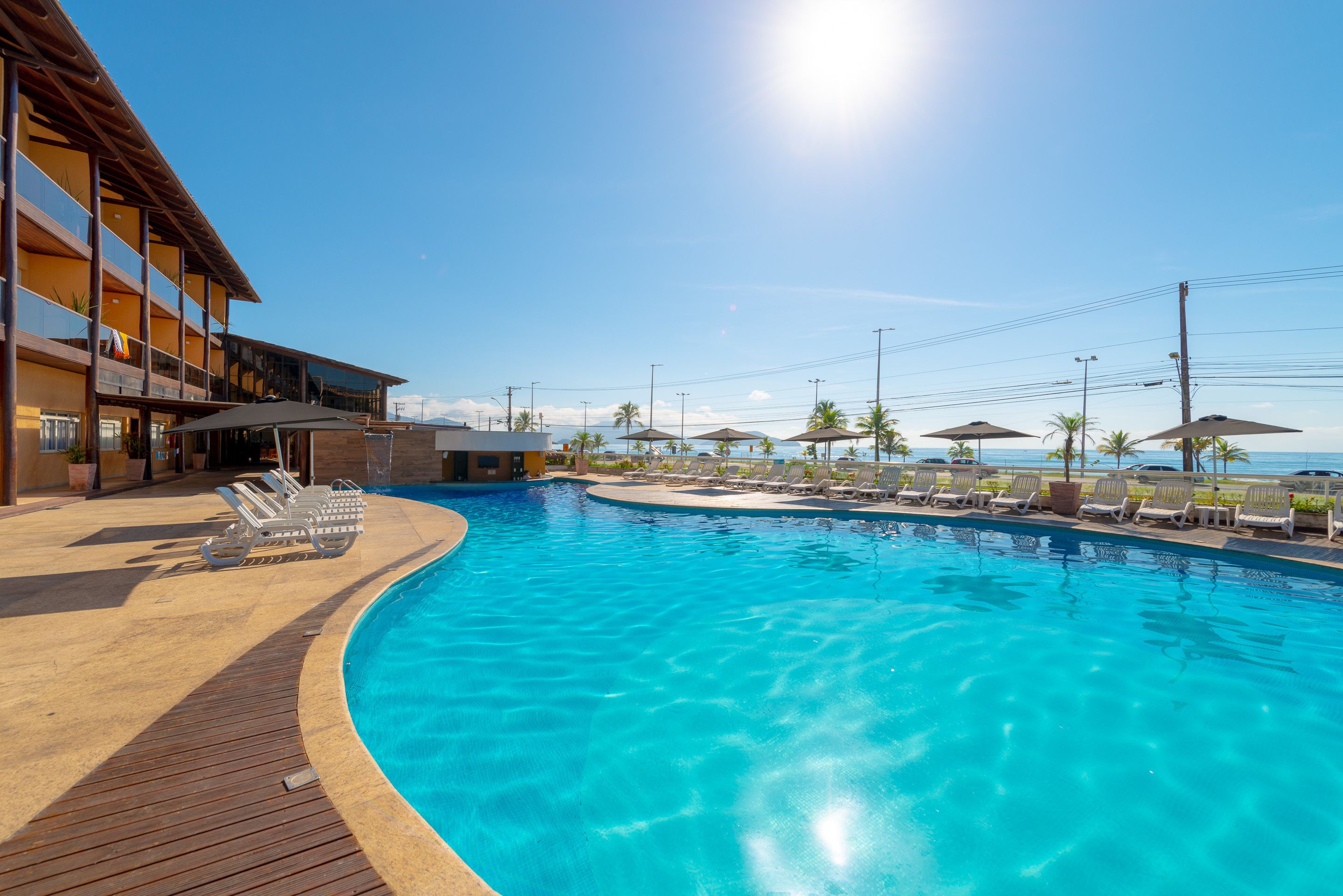 A swimming pool with lounge chairs and umbrellas, surrounded by a building with multiple floors and a clear sky.