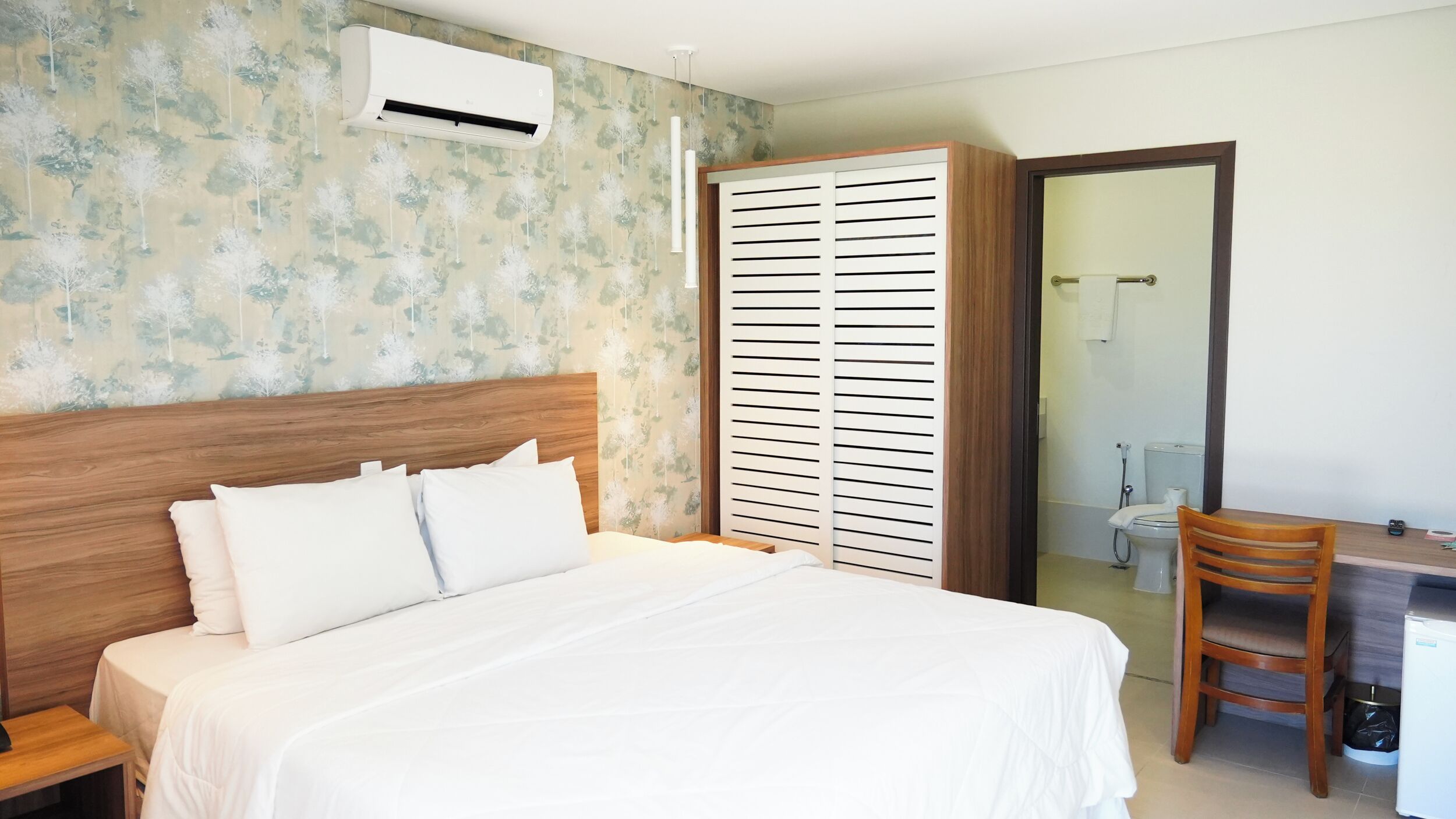 Un dormitorio con cabecera de madera, cama con ropa de cama blanca, armario con listones horizontales, escritorio con silla, y un baño visible a través de una puerta abierta.