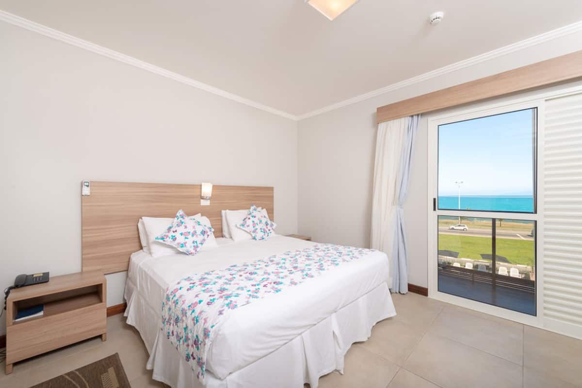 A bedroom with a large bed, a wooden headboard, a nightstand, a view of the sea through a balcony window.