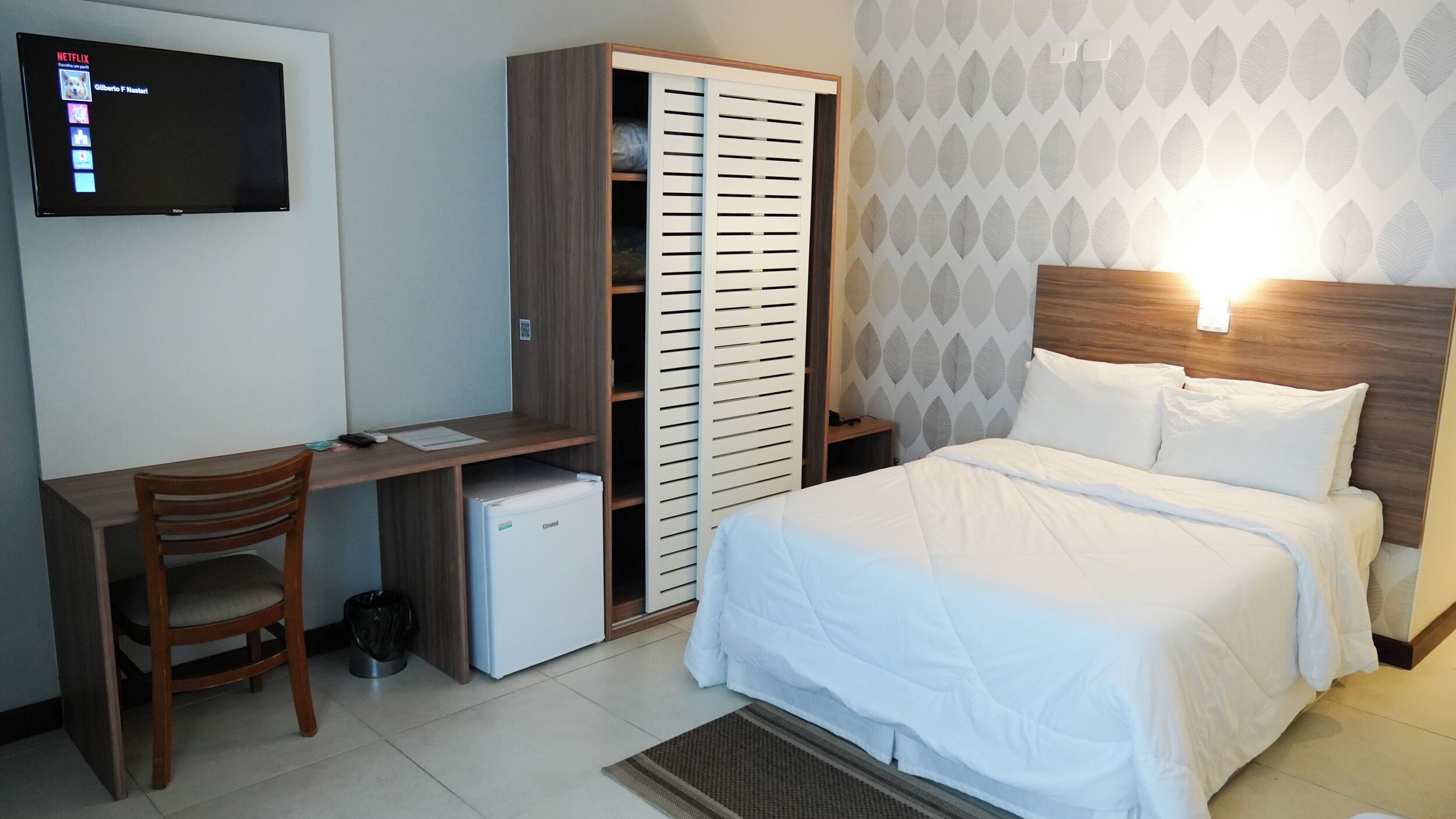 A hotel room with a bed, a desk with a chair, a wardrobe, a television mounted on the wall, and a patterned wallpaper.