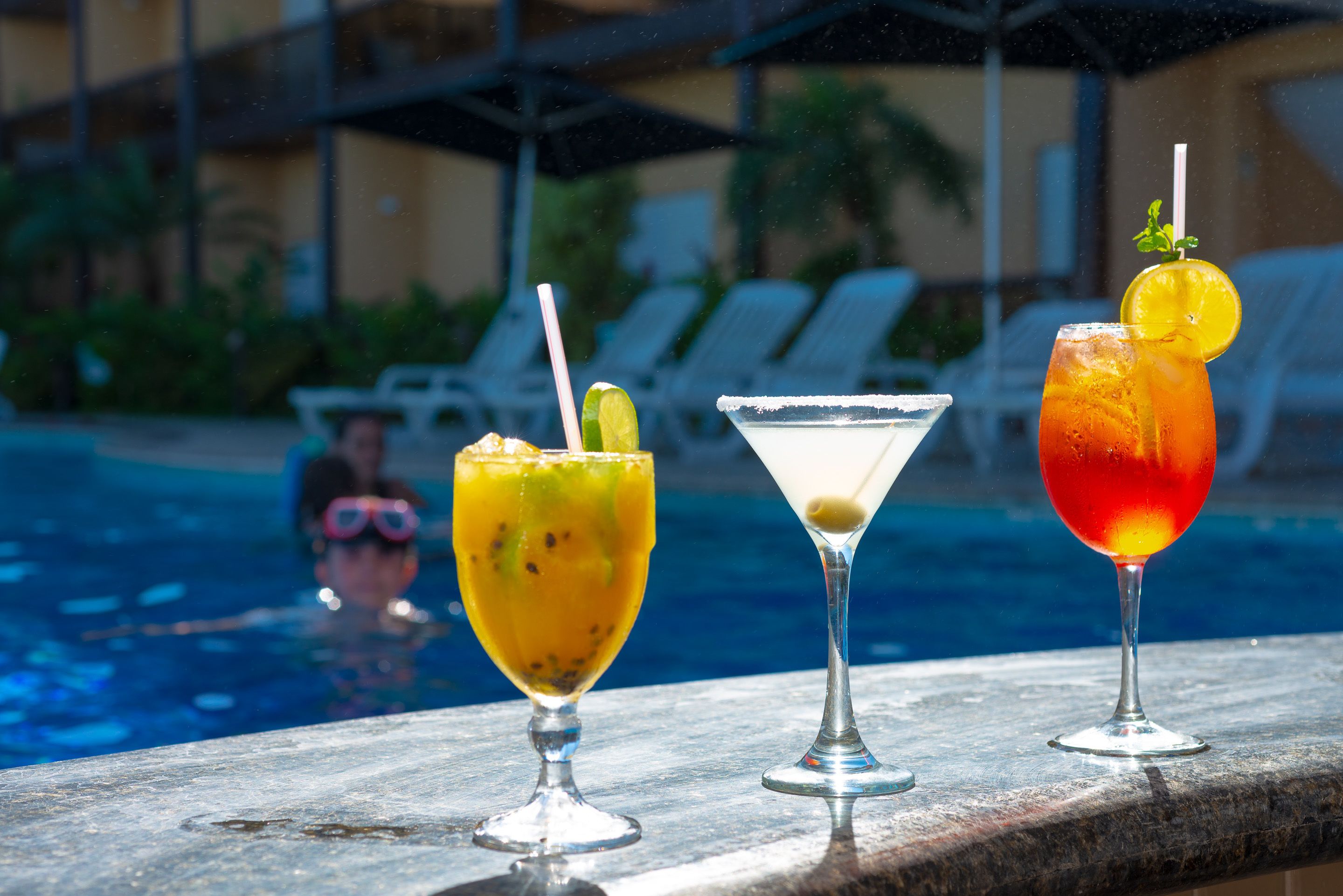 Three cocktails on a poolside table with a pool and lounge chairs in the background.