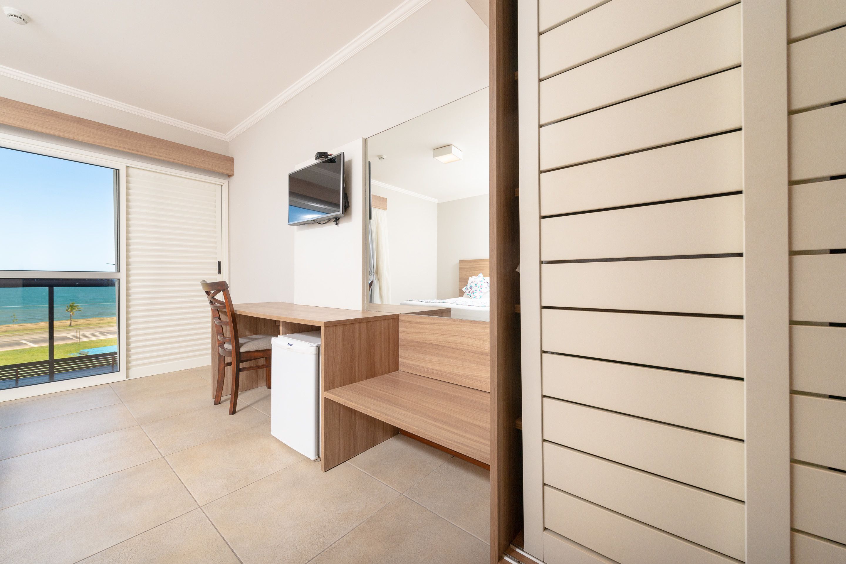 Ein modernes Schlafzimmer mit Schreibtisch aus Holz, einem an der Wand montierten Fernseher und einem großen Fenster mit Blick auf den Strand.