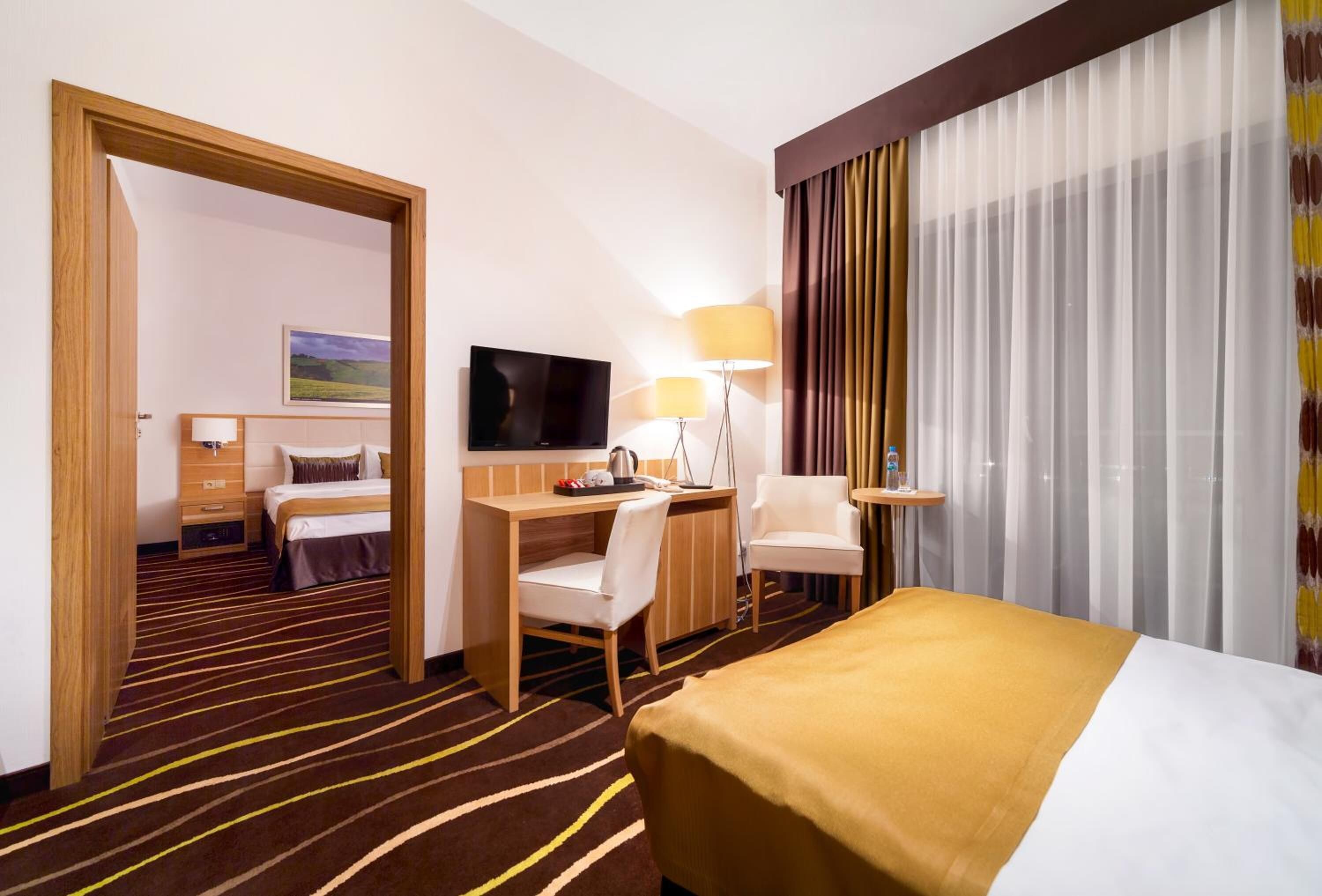 Quarto de hotel com cama, escrivaninha, cadeira, televisão e abajur.