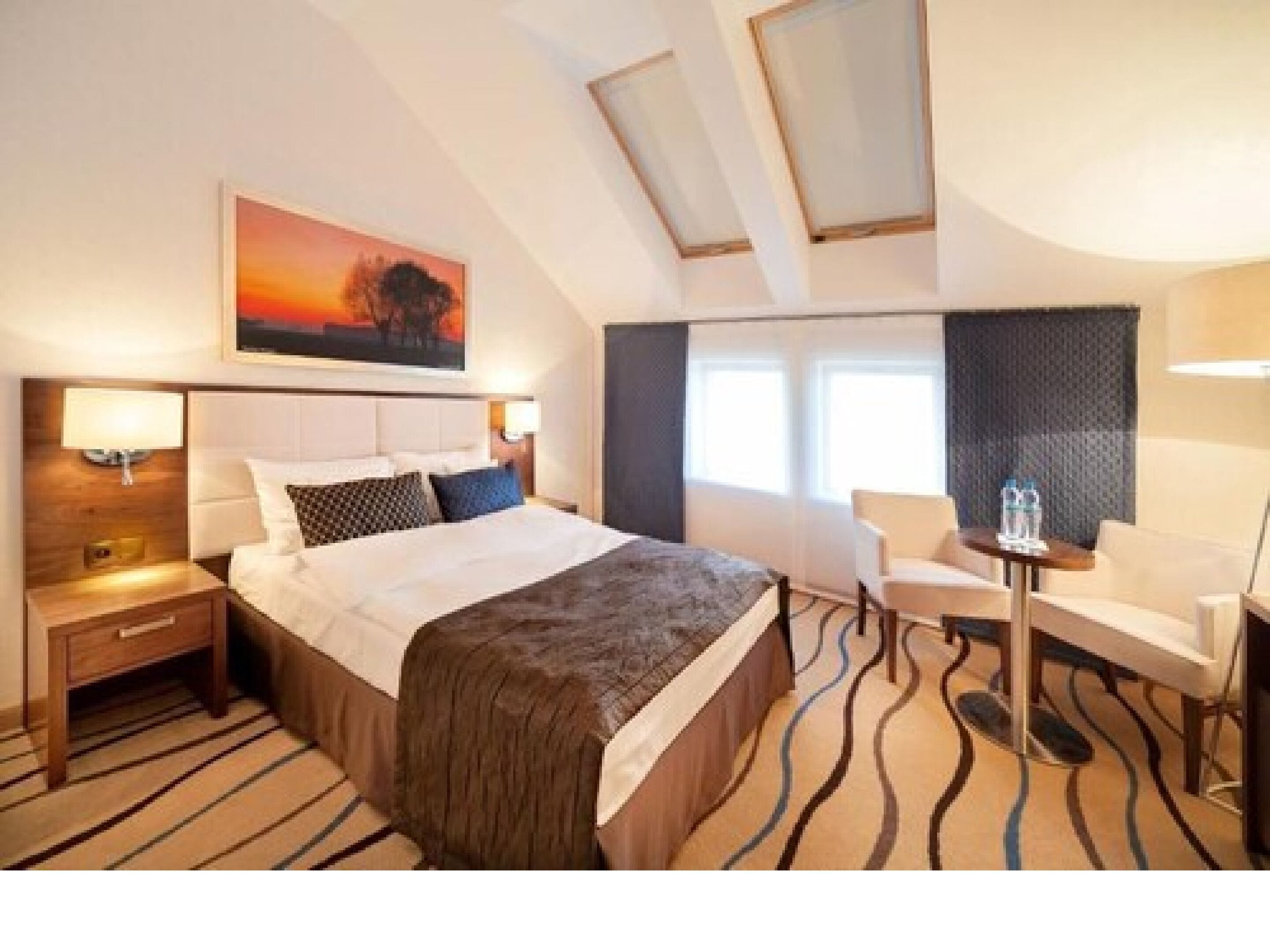 Hotelkamer met een groot bed, een bureau en een stoel. Er hangt een schilderij aan de muur en er is een raam met gordijnen.