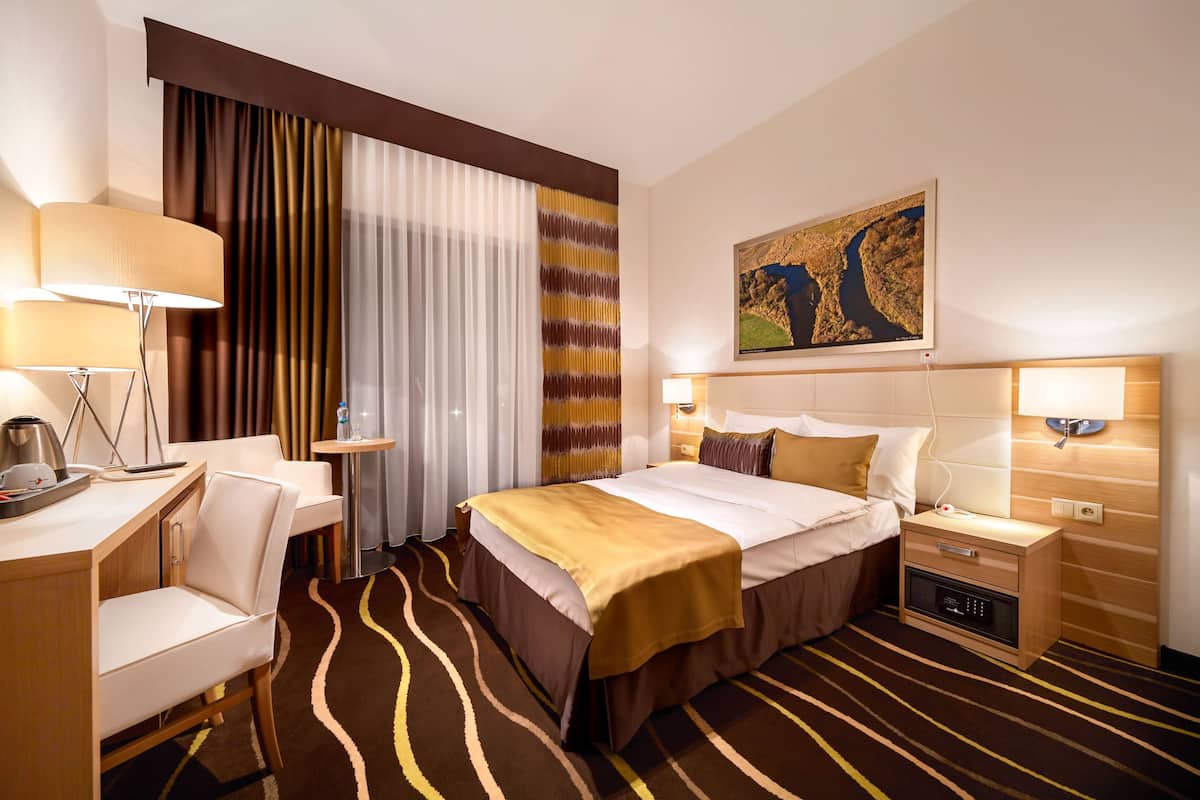 Ein Hotelzimmer mit Bett, Nachttischen, Schreibtisch, Stuhl, einem großen Fenster mit Vorhängen und einem Bild an der Wand.