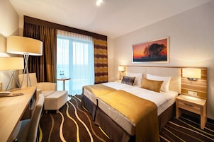 Quarto de hotel com cama, mesas de cabeceira, escrivaninha, cadeira, mesa pequena e janela com cortinas.