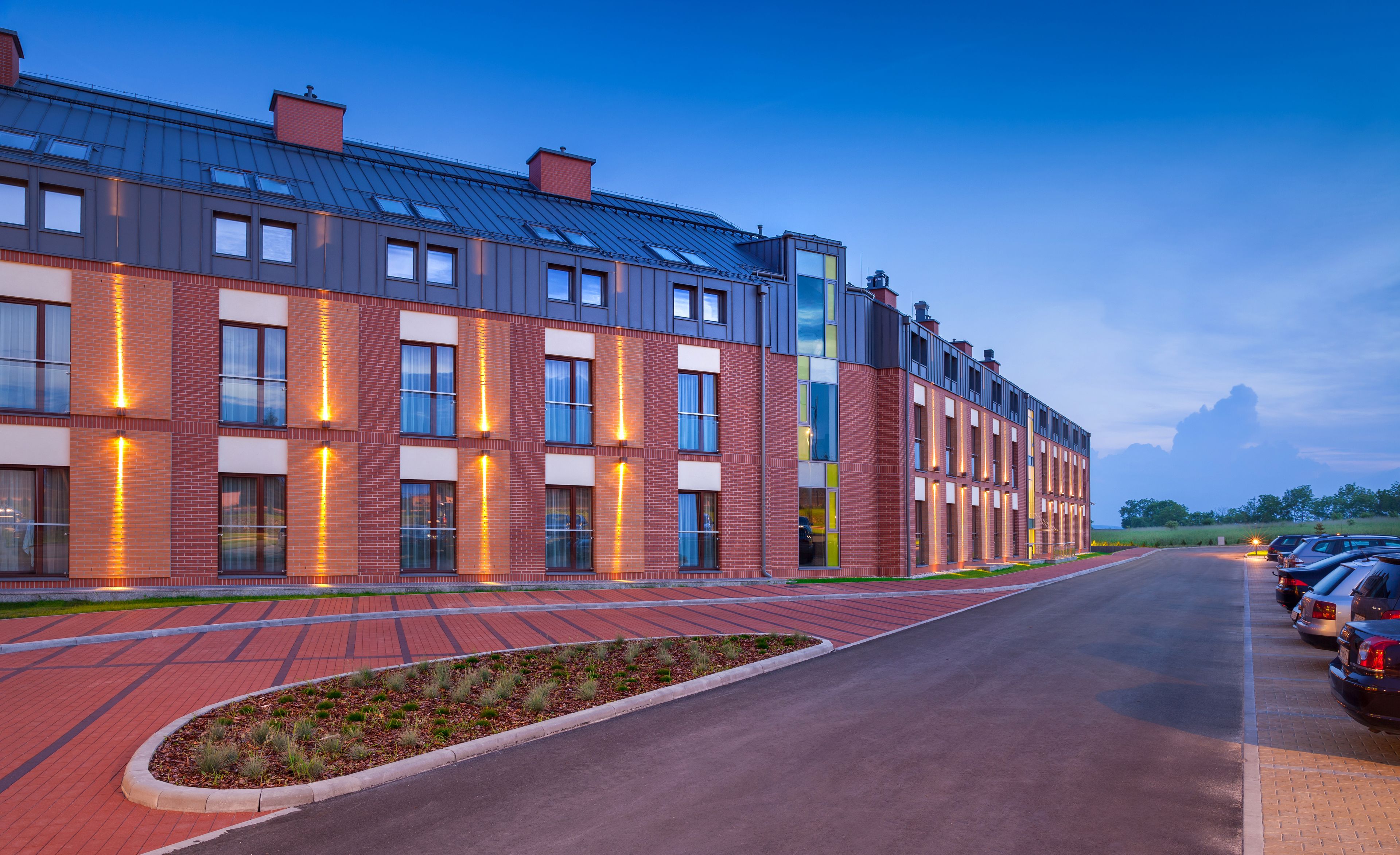 Un bâtiment moderne avec une façade en briques, de grandes fenêtres et un espace de stationnement où sont garées des voitures.