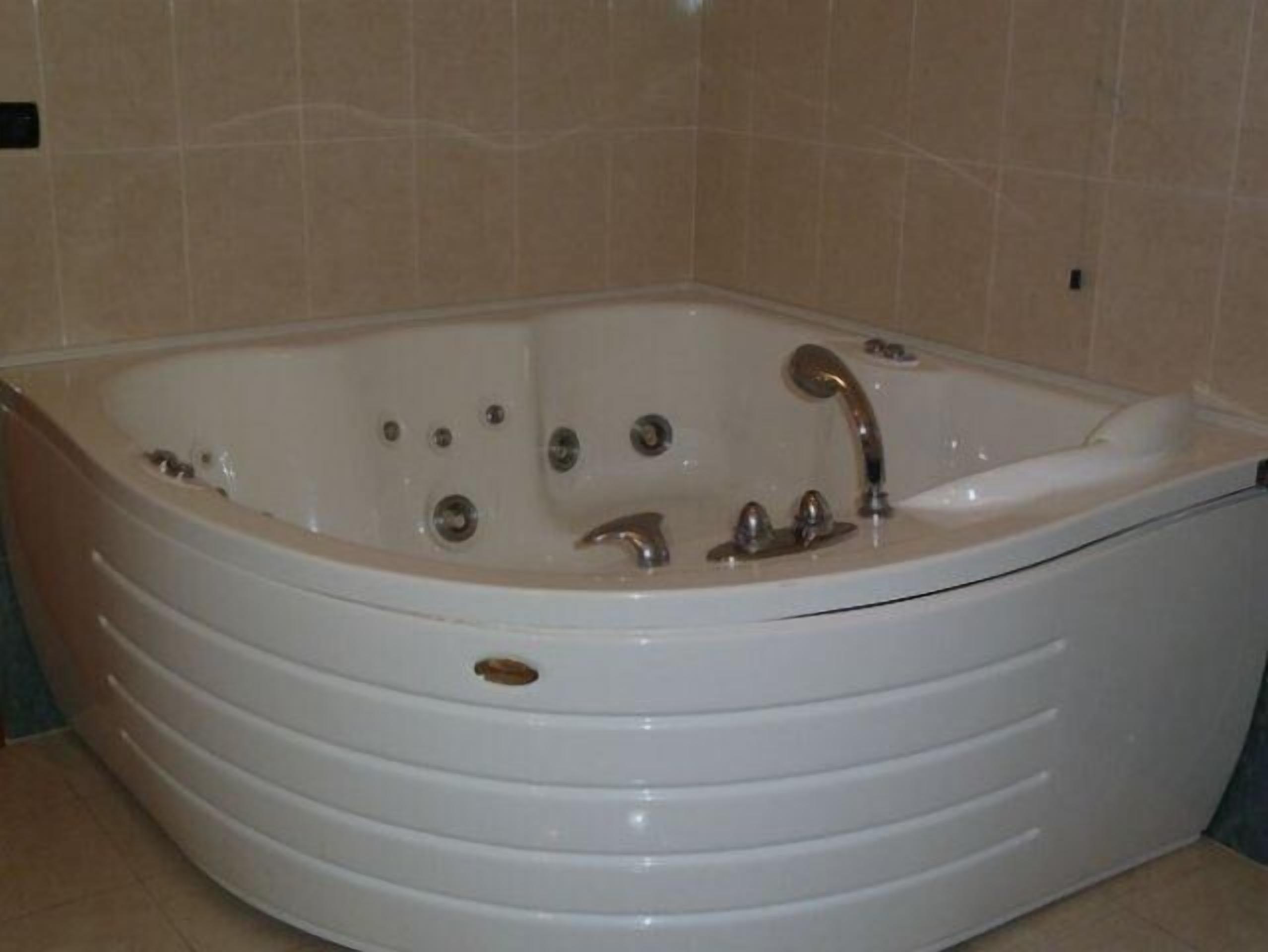 A large, white, freestanding bathtub with multiple faucets and jets, set against beige tiled walls.