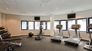 Un gimnasio con cintas de correr, elípticas, una prensa de banca y un estante con mancuernas.