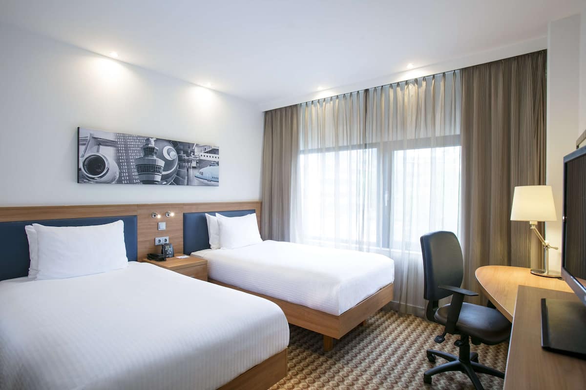 Quarto de hotel com duas camas, uma escrivaninha, uma cadeira, um abajur e uma janela grande com cortinas.