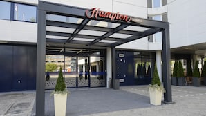 Entrada de un hotel con un corredor cubierto, un letrero que dice "Hampton by Hilton" y plantas en macetas.