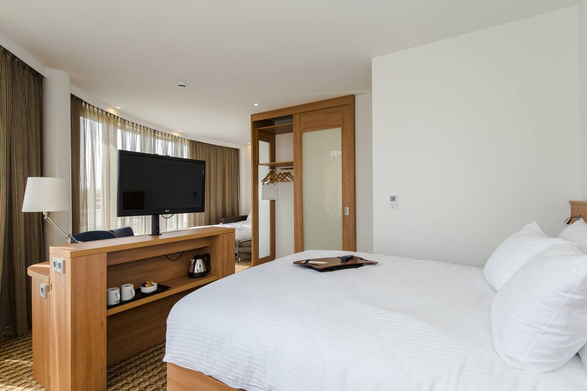 Quarto de hotel com uma cama grande, uma mesa de trabalho com televisão de tela plana, um guarda-roupa e um espaço de estar com luminária.