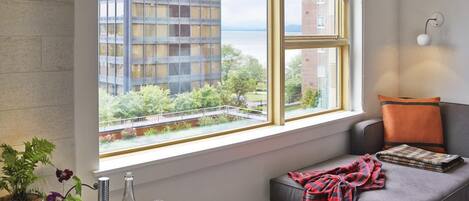 A modern living room with a large window, a grey sofa, a small table with a bottle and glasses, and a view of a building across the water.