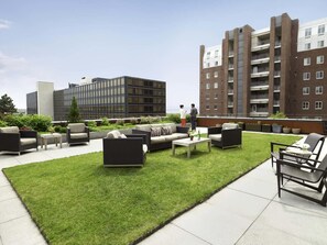 A rooftop patio with outdoor furniture, a grassy area, and a view of a modern building and residential apartments. - Hotel Vermont (Burlington)
