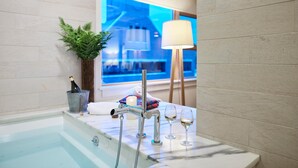 Un baño moderno con bañera de mármol, cabina de ducha de vidrio, una planta en maceta, una lámpara y una ventana con vistas a un edificio.