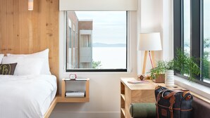 Una habitación de hotel moderna con cabecera de madera, cama con ropa blanca, mesita de noche con lámpara y planta, escritorio con computadora portátil y bolso, y ventana con vistas a edificios y agua.