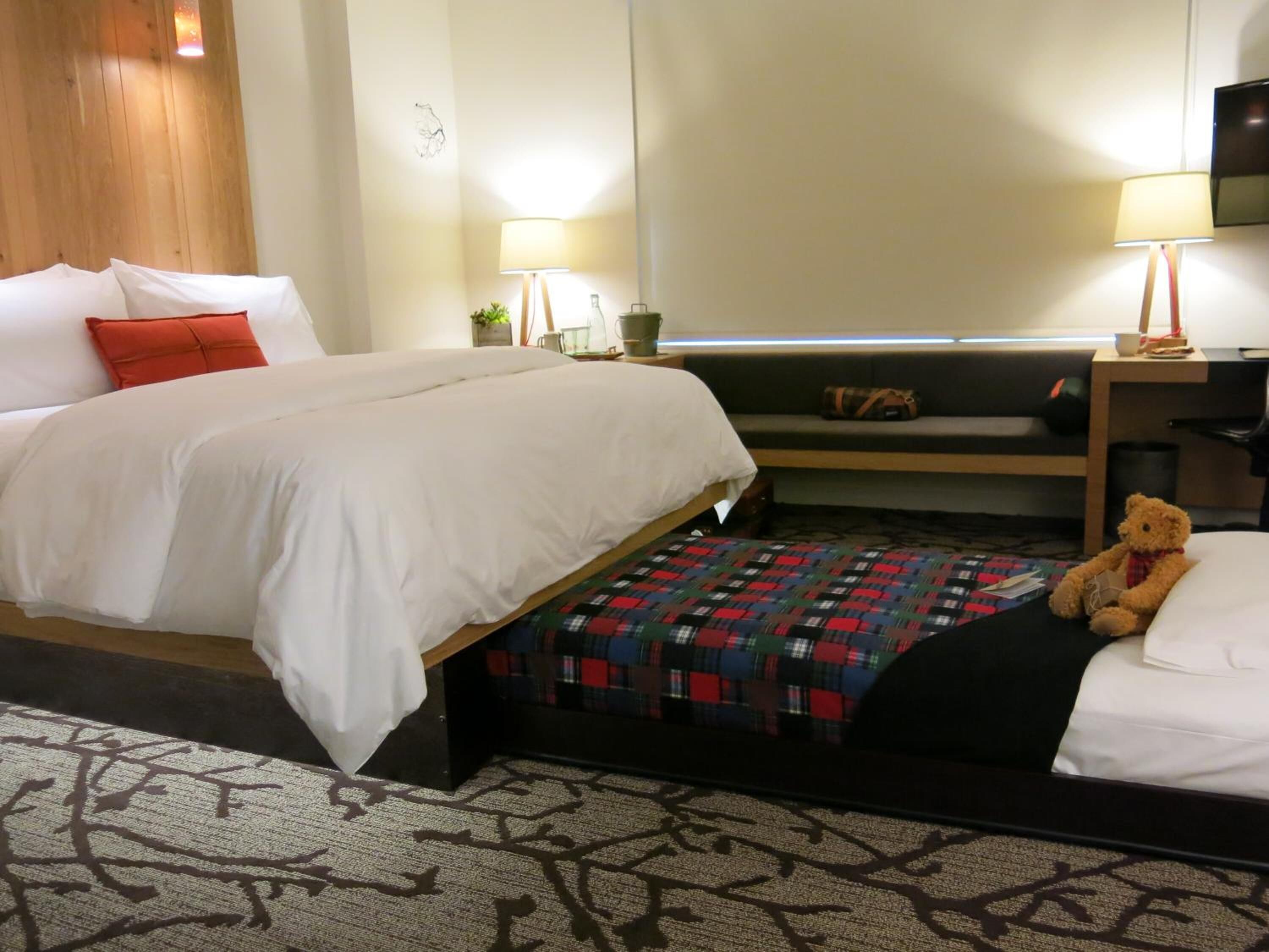 Quarto de hotel com duas camas, um sofá, uma TV e um urso de pelúcia.