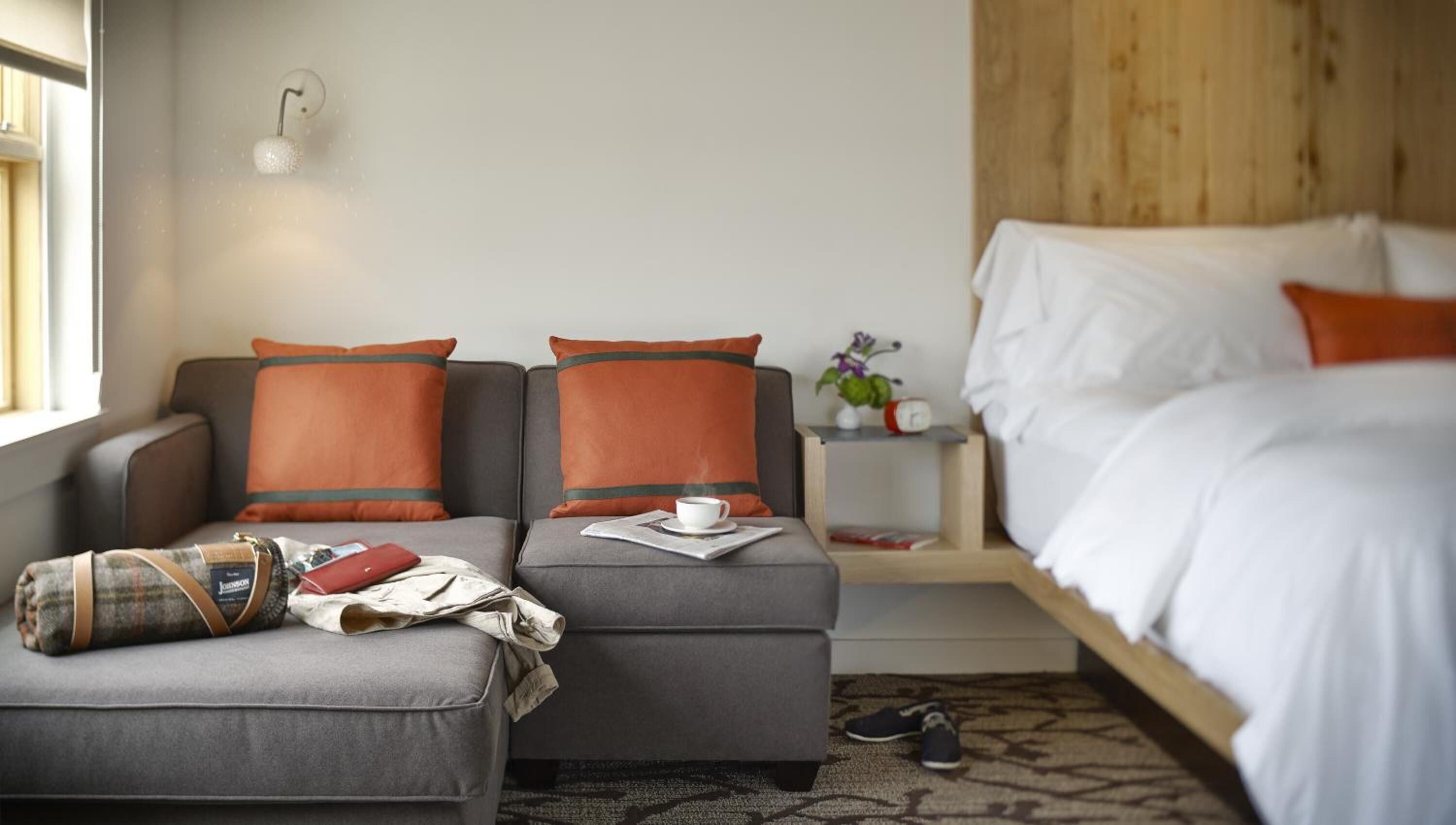 Hotelkamer met een grijze bank, twee oranje kussens, een klein tafeltje met een kop en krant, een bed met houten hoofdbord en een nachtkastje met klok en plant.