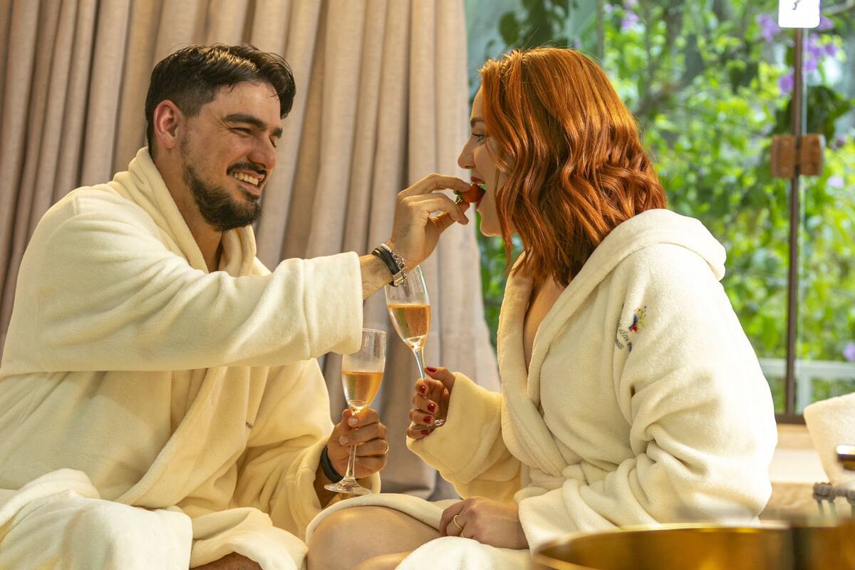 Een koppel in badjassen proost met champagnefluiten in een kamer met een groot raam, een bad en een zichtbare plant buiten.
