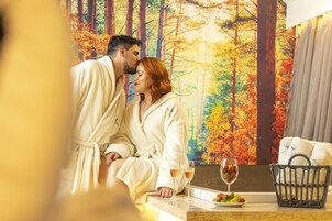 Um casal de roupão senta-se junto a uma janela com vista para uma floresta, com uma cesta de frutas e duas taças de vinho sobre a mesa.