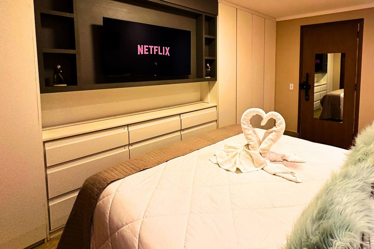 Een hotelkamer met een grote flatscreen-tv waarop 'NETFLIX' staat, een netjes opgemaakt bed met een hartvormig kussen en een zachte witte plaid.