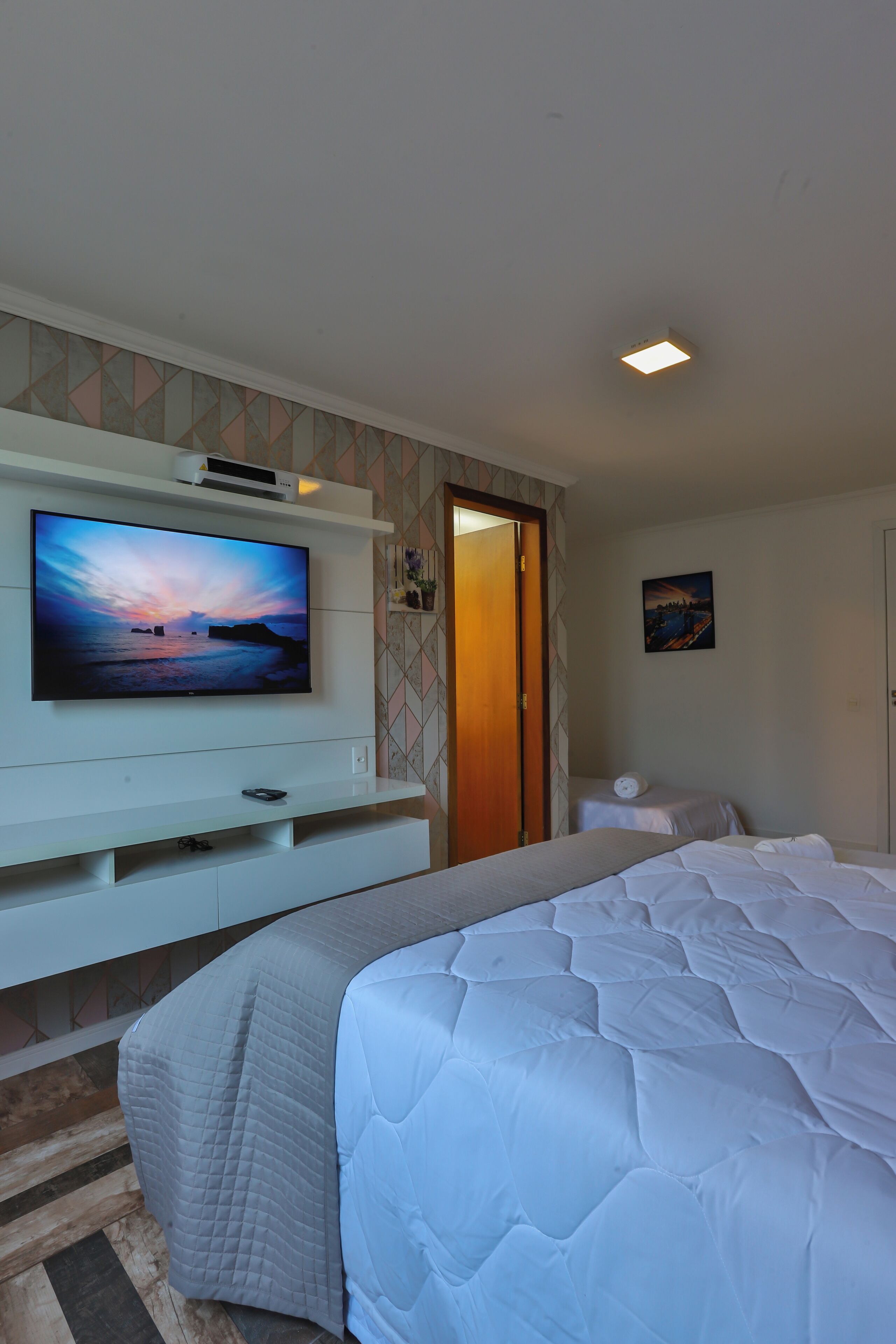 A hotel room with a large bed, a television mounted on the wall, a bedside table, and a door leading to another room.