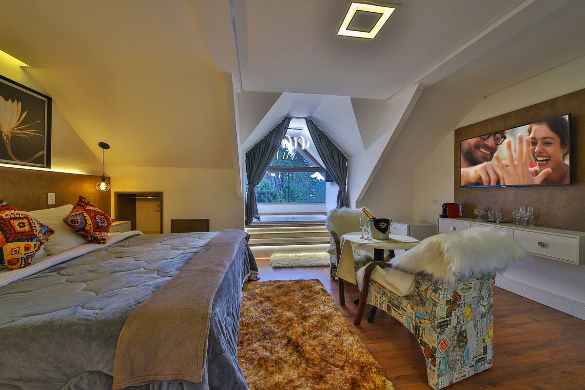 Een slaapkamer met een groot bed, een klein tafeltje met een stoel, een televisie en uitzicht op de omgeving door een raam.