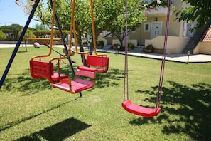 A playground with swings and a slide, surrounded by a well-maintained lawn and a building with a tiled roof. - Villa Carina (Kefalonia)