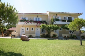 A two-story building with a yellow facade, white shutters, and balconies with pink flowers. - Villa Carina (Kefalonia)
