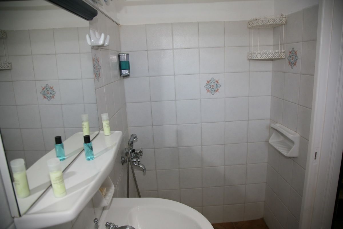 A bathroom with a white sink, mirror, and tiled walls. There are toiletries on the sink and a shower area with a white shelf.