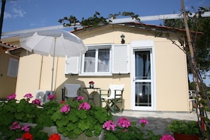 A building with a beige exterior, white shutters, and a white umbrella. There are potted plants with pink flowers in front. - Villa Carina (Kefalonia)