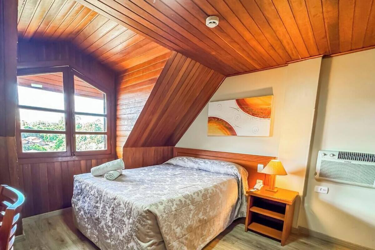 Un dormitorio con techo inclinado, una cama con colcha estampada, una mesita de noche con lámpara, una ventana con vista a follaje y un aire acondicionado instalado en la pared.