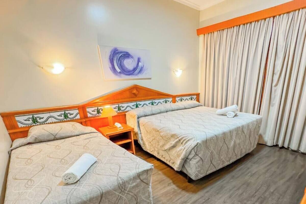 Habitación de hotel con dos camas, una mesita de noche, una lámpara, un cuadro en la pared y un ventanal con cortinas.