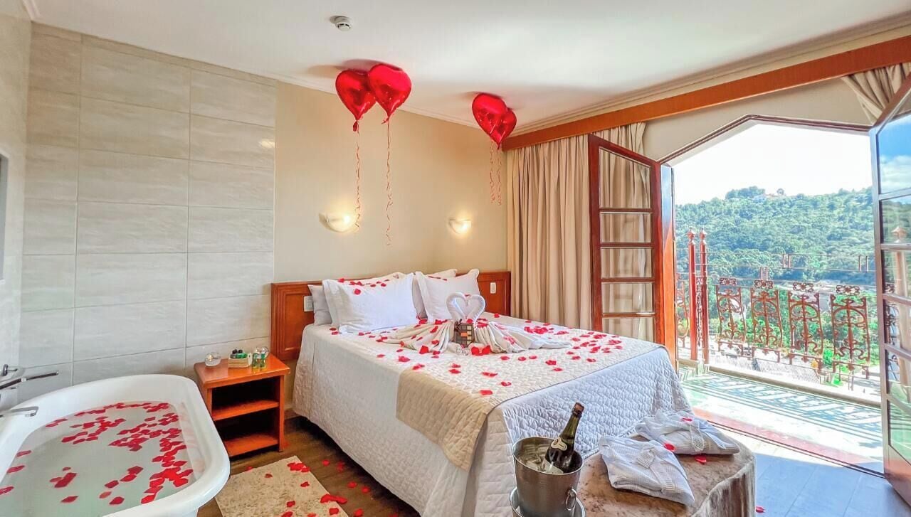 A hotel room with a bed, a bathtub with rose petals, a champagne bottle, and a balcony with a view.