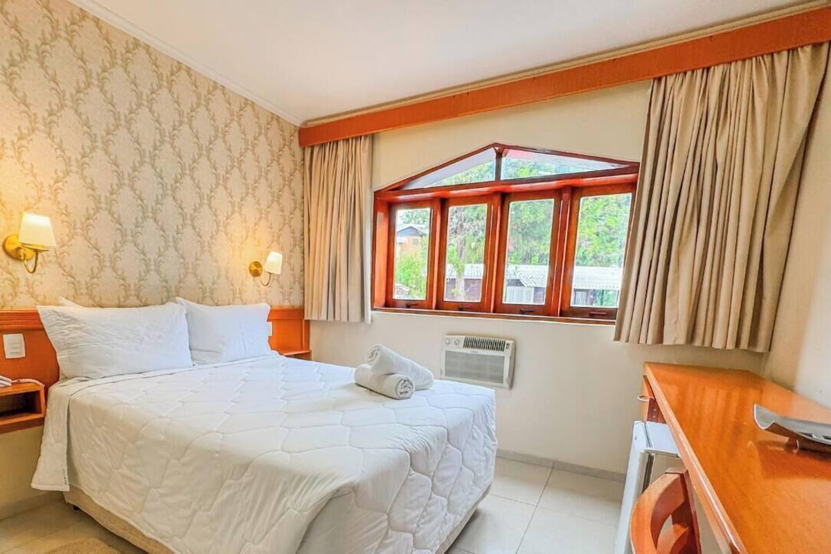 Una habitación de hotel con una cama grande, dos lámparas de noche, un ventanal con cortinas, un escritorio de madera y un radiador.